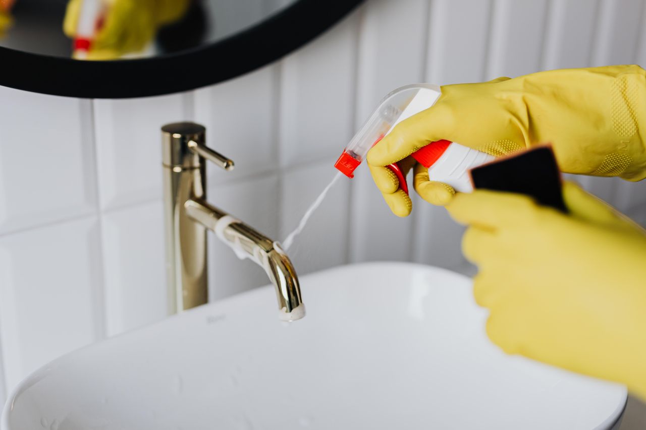 Seen from above, an anonymous housekeeper in yellow rubber gloves sprays cleaner toward a chrome water faucet in a bathroom